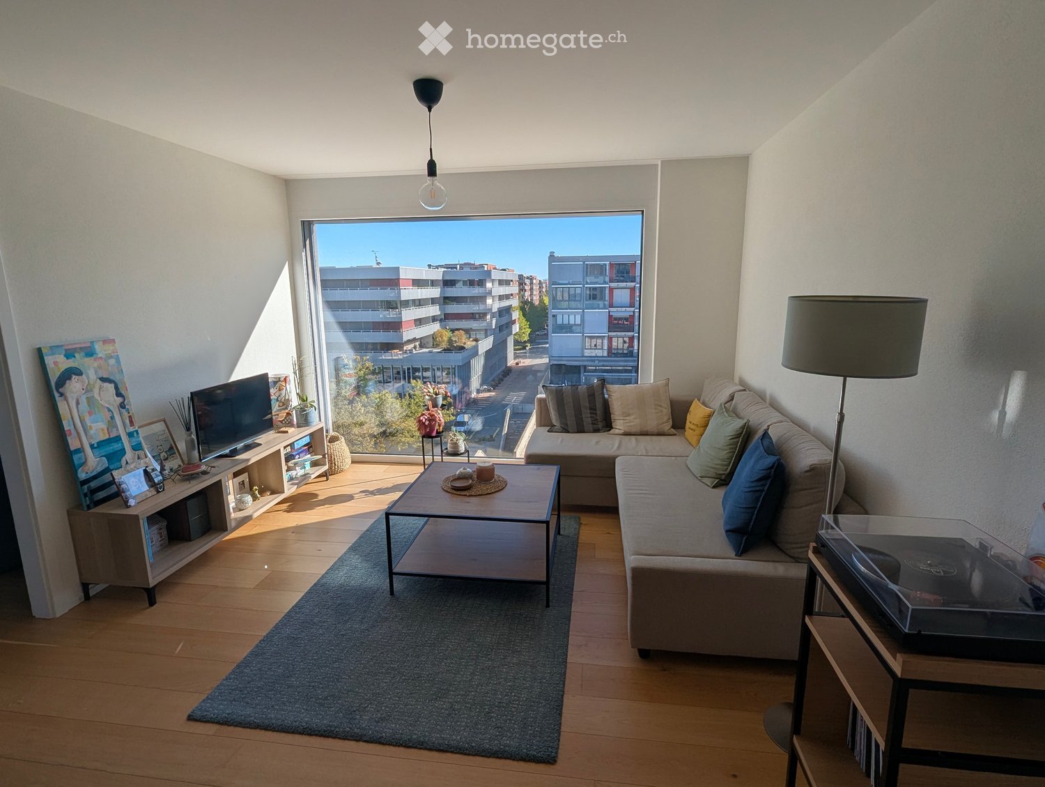 Modern living room with large windows offering a city view, featuring a grey sofa with multiple cushions, a wooden coffee table, a floor lamp, a TV stand with a flat-screen TV, and a painting on the wall.