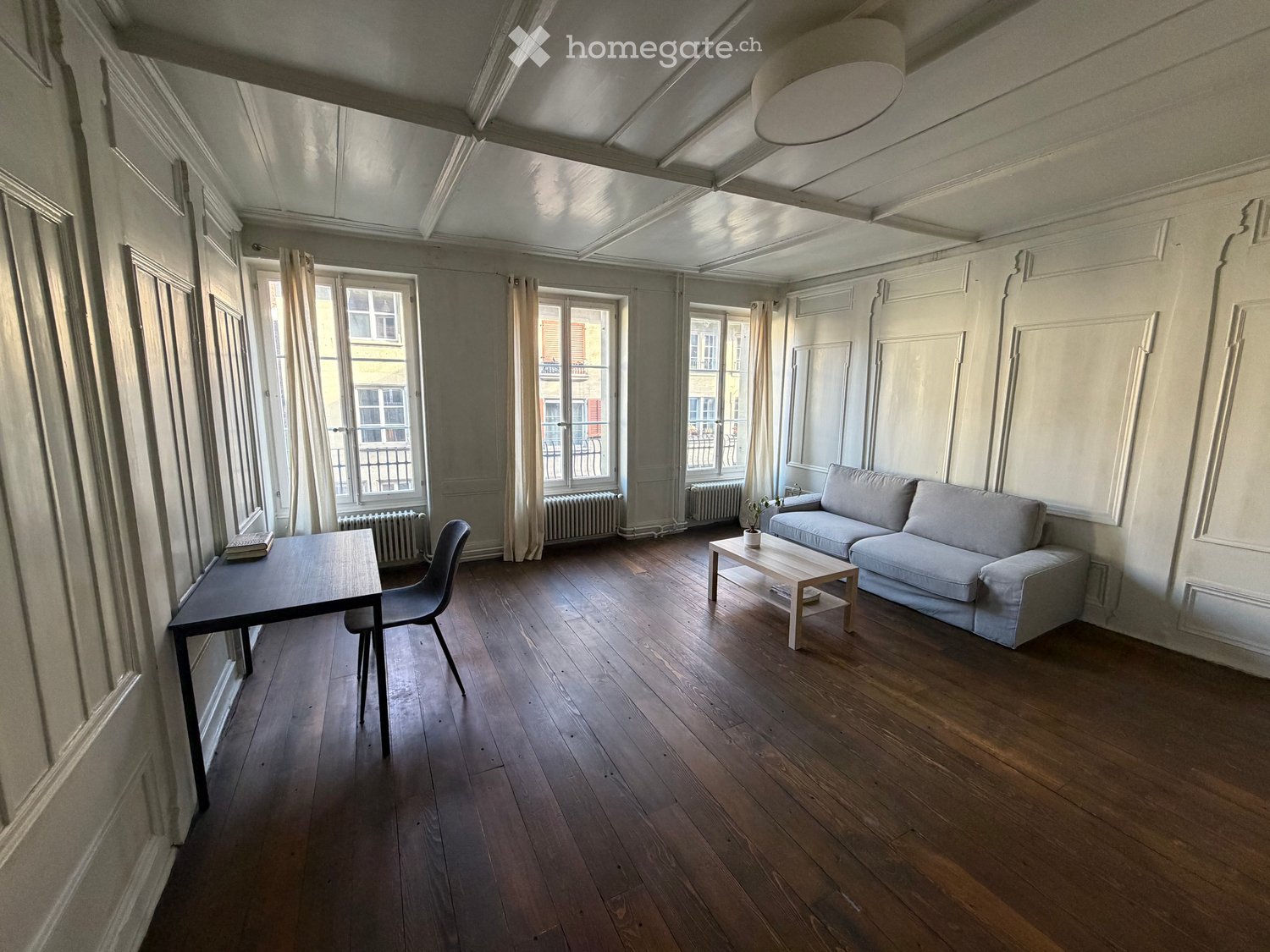 Living room with wooden flooring, large windows, a sofa, coffee table, and a small table with a chair.
