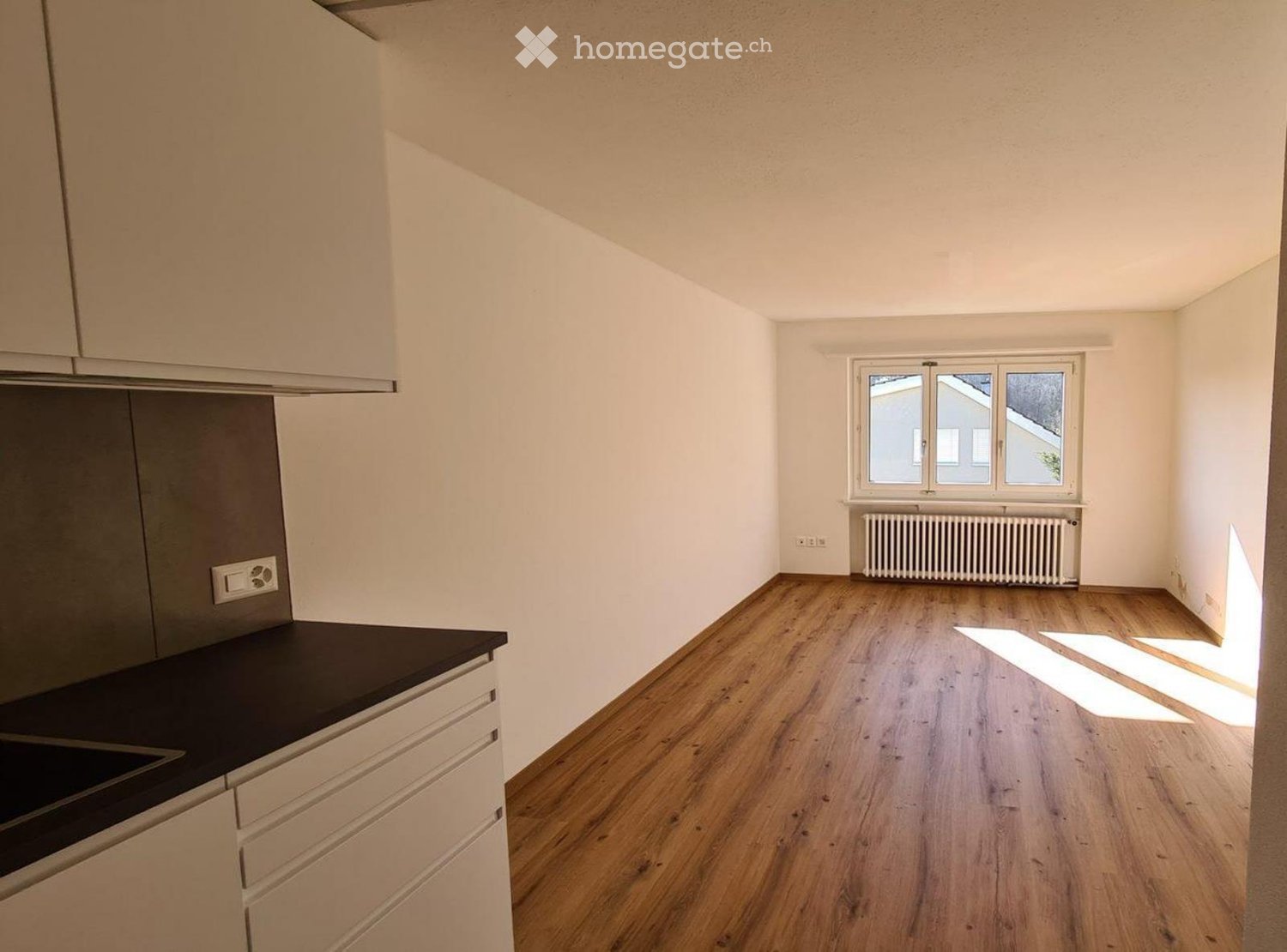 Empty kitchen with light-colored cabinets, dark countertop, wooden flooring, and a window with white frame and radiator below.