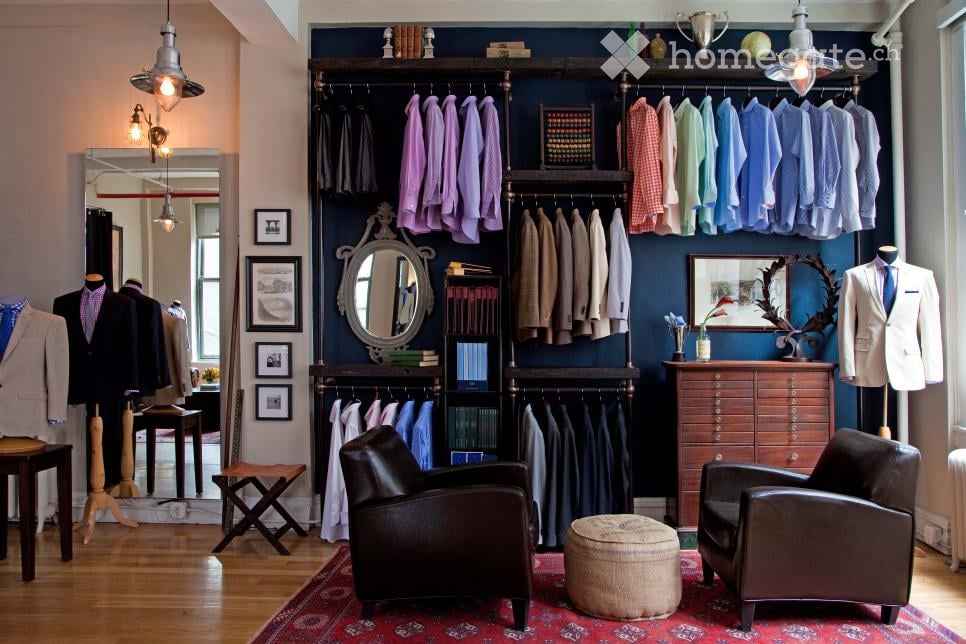 A stylishly furnished room with a dark blue accent wall, wooden floor, and a variety of clothing items displayed on racks and mannequins. The room features leather chairs, a round ottoman, a small wooden table, and several framed pictures on the wall.