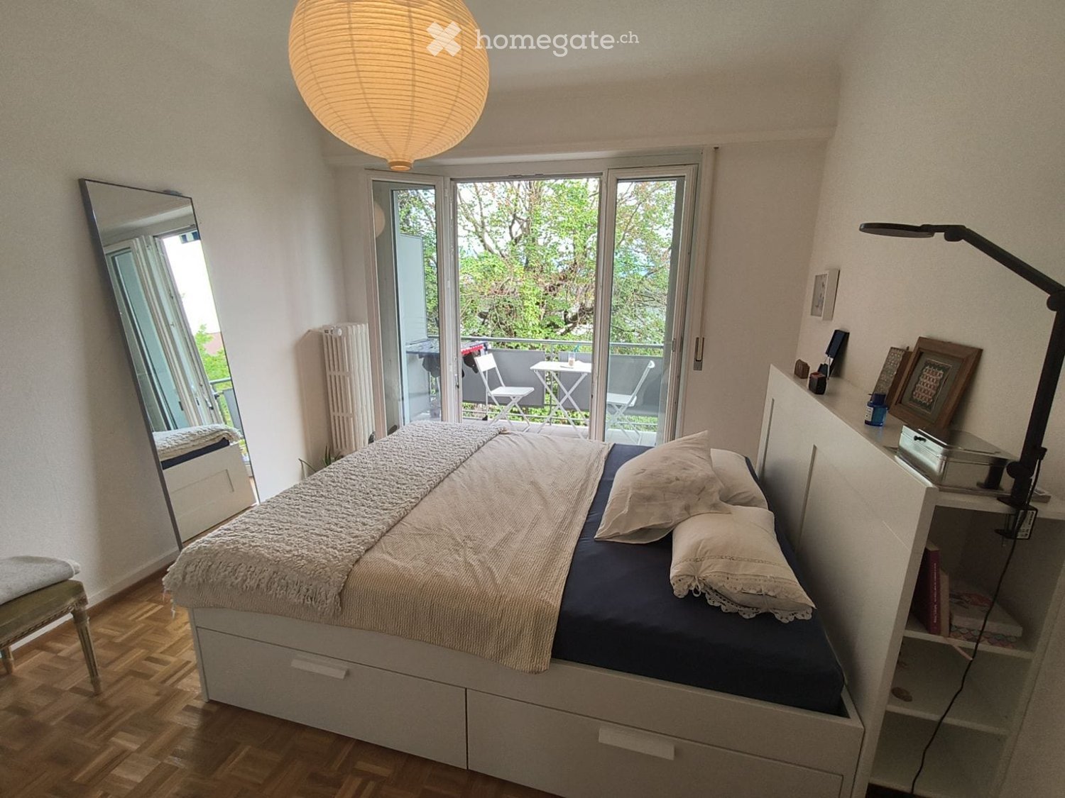 Cozy bedroom with a single bed, nightstand, mirror, and a balcony view. The room features wooden flooring and a large window leading to a balcony with outdoor furniture.