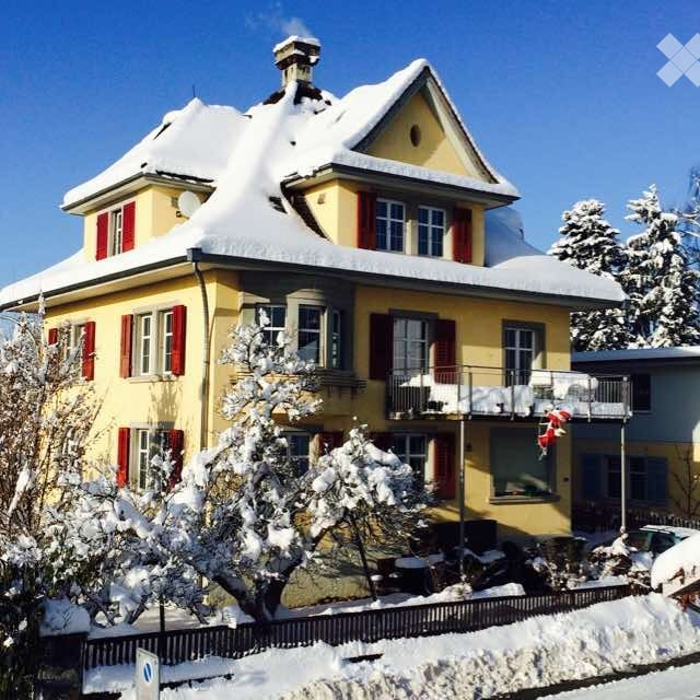 A two-story yellow house with red shutters and snow-covered roof and balcony. The house features multiple windows and a chimney, surrounded by snow-covered trees and a fence.