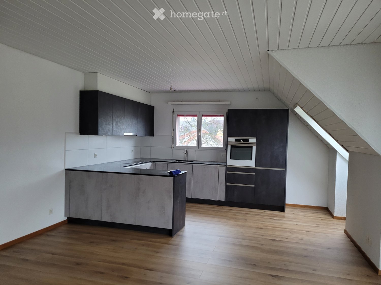 Modern kitchen with grey cabinets, a central island, and a wooden floor. Features include a sink, oven, and ample natural light from a large window.