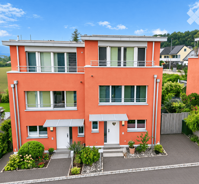Two-story house with orange exterior, white-framed windows, and a white front door. Features a small garden with shrubs and potted plants, and a paved driveway.