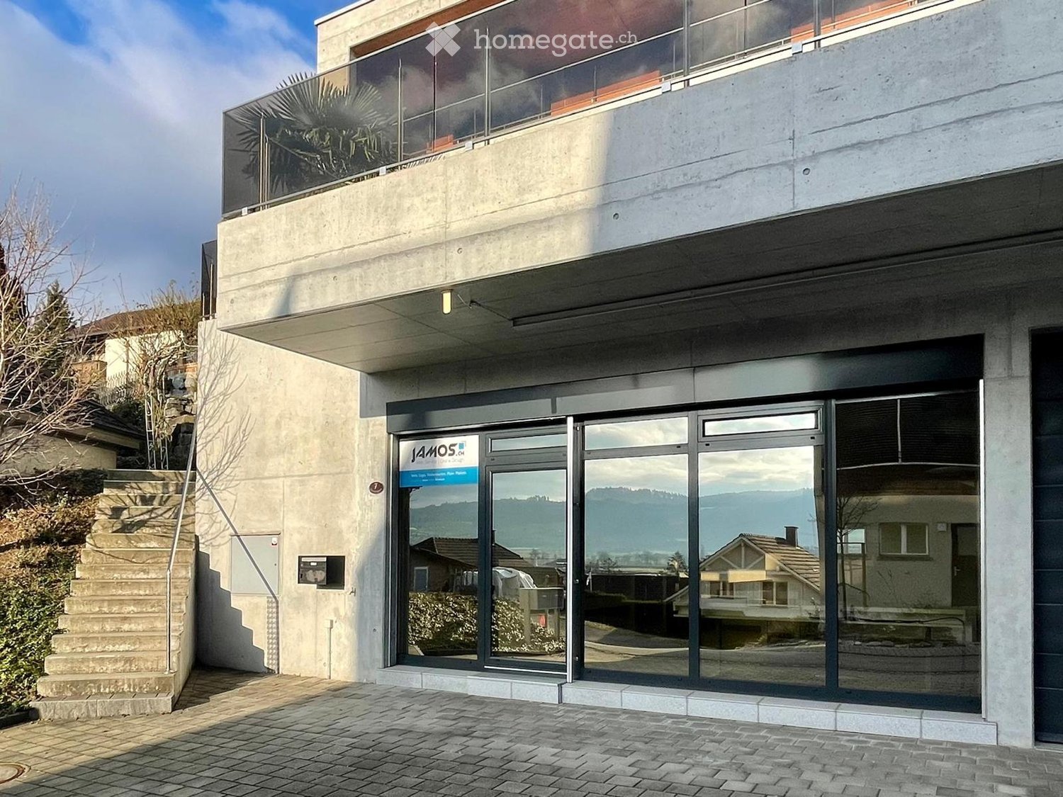Modern two-story building with a glass entrance, concrete facade, and a raised ground floor accessed by stairs.