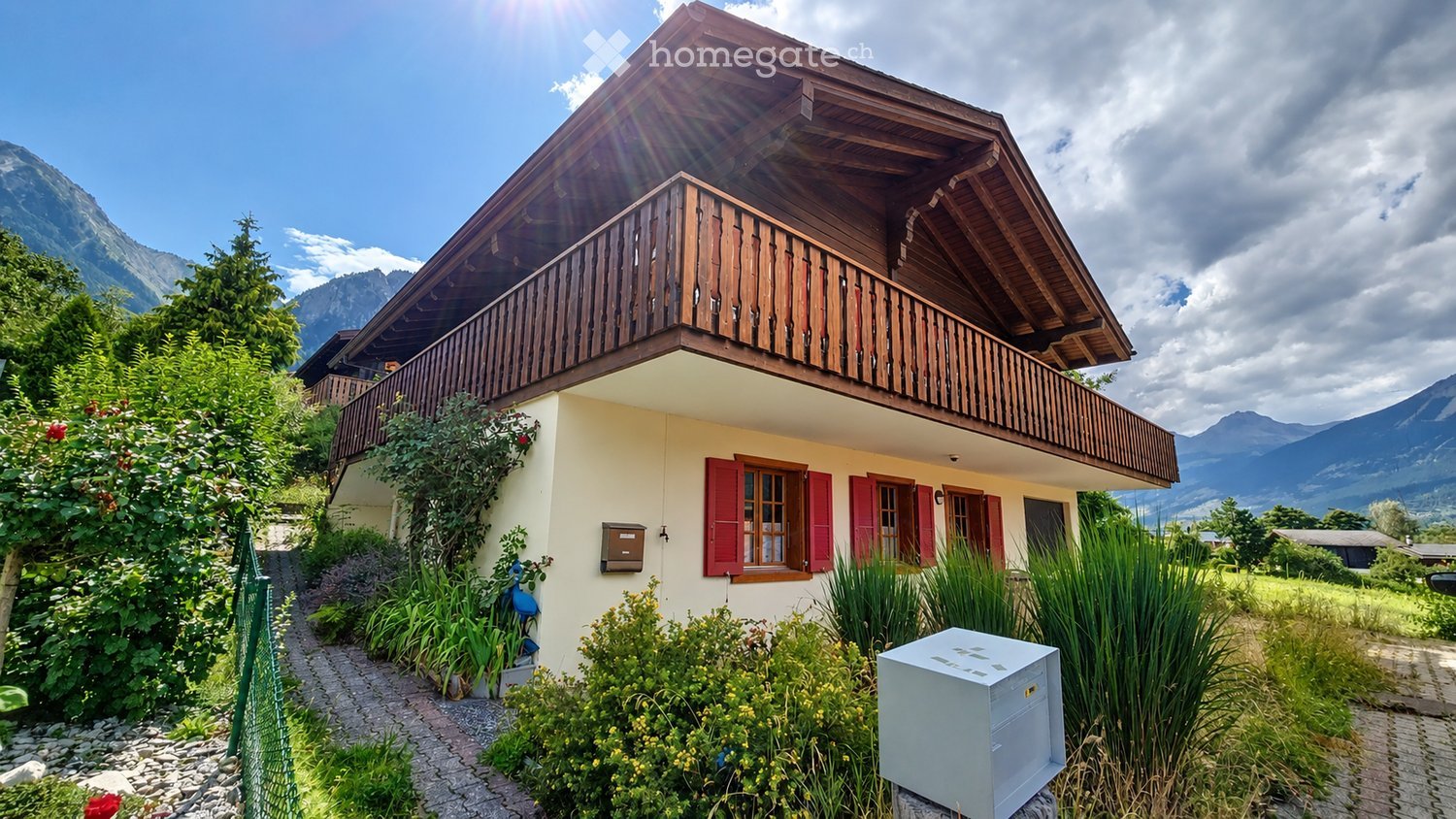 Two-story wooden house with a balcony, red shutters, and a view of mountains. The house is surrounded by greenery and a well-maintained garden.