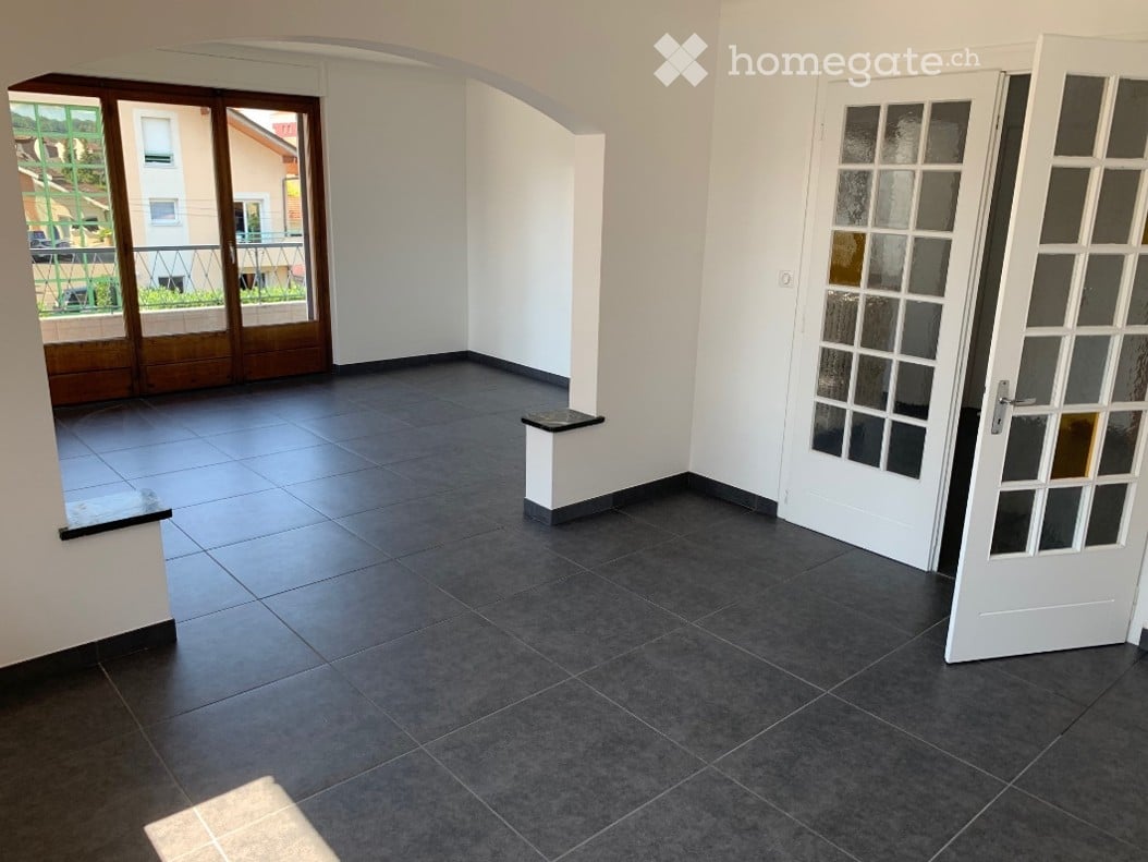 Empty room with dark grey tiled floor, white walls, and a small white bench. Features a large sliding glass door leading to a balcony and a white door with glass panels.