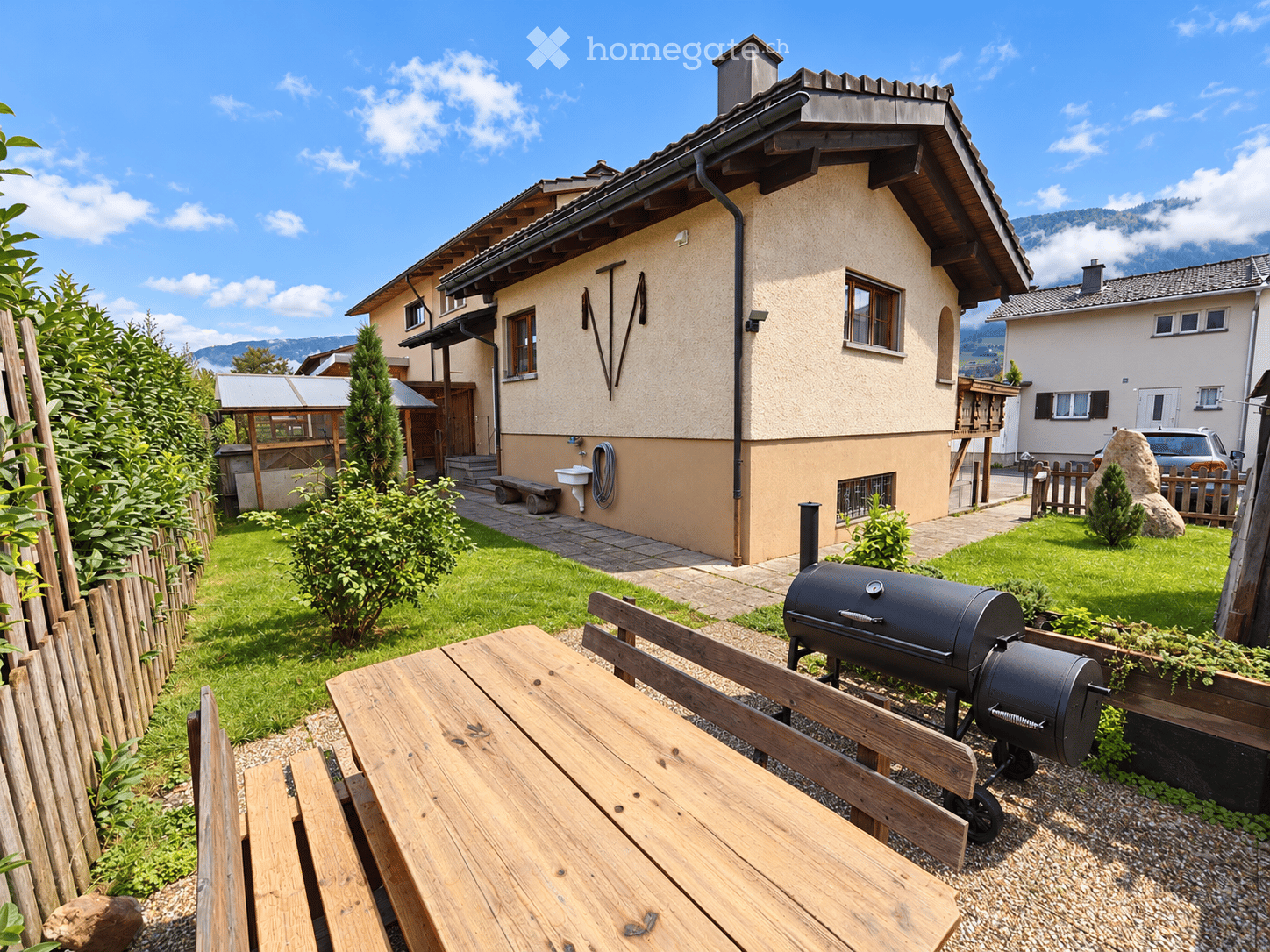 A well-maintained garden area with a wooden table, benches, and a grill, surrounded by greenery and a wooden fence, with a house and mountains in the background.