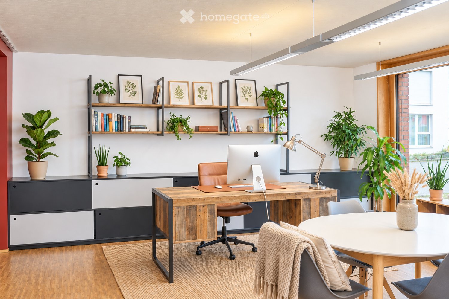 Modern office space with a wooden desk, ergonomic chair, laptop, floor lamp, shelves with books and plants, and a round meeting table with chairs.