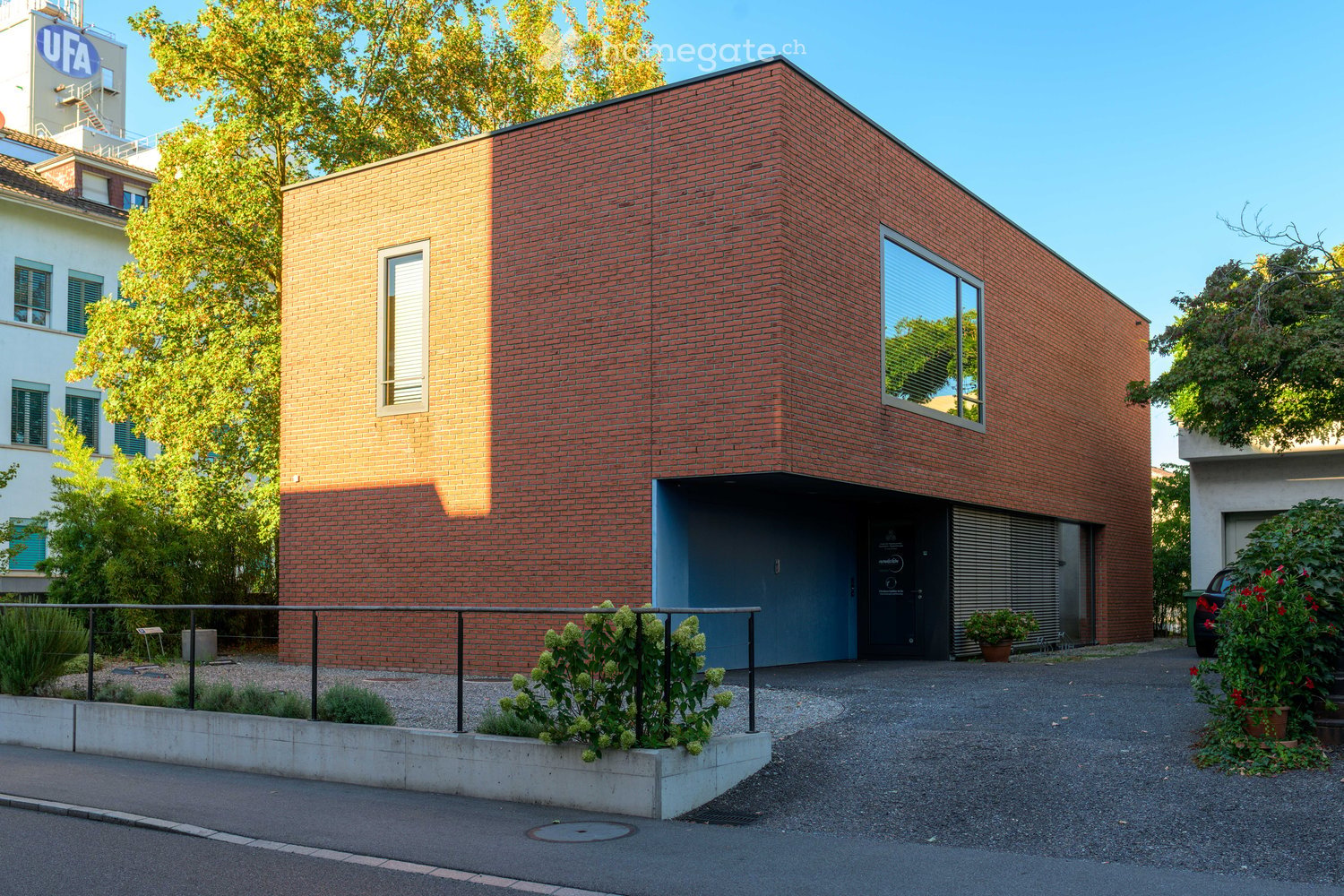 Modern red brick building with a flat roof, large windows, and a blue garage entrance. The building is surrounded by greenery and a paved driveway.