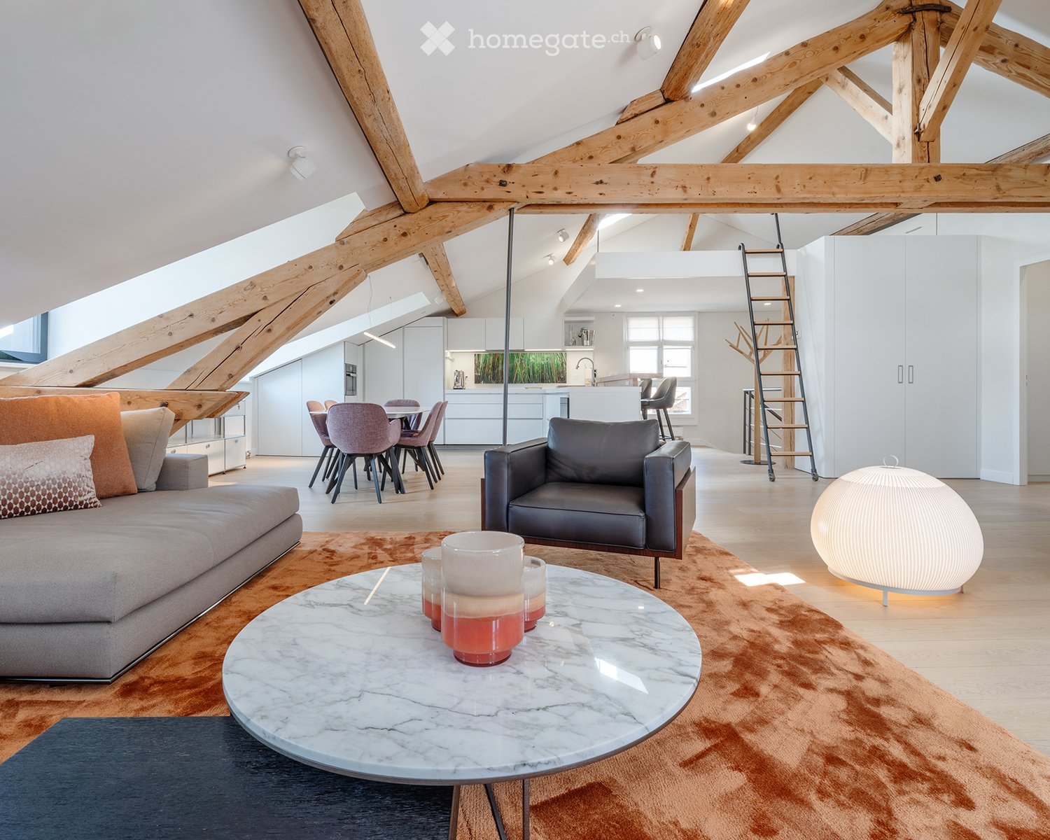 Modern living room with exposed wooden beams, a grey sofa, a round marble coffee table, an armchair, a dining area with chairs, and a round ball lamp.