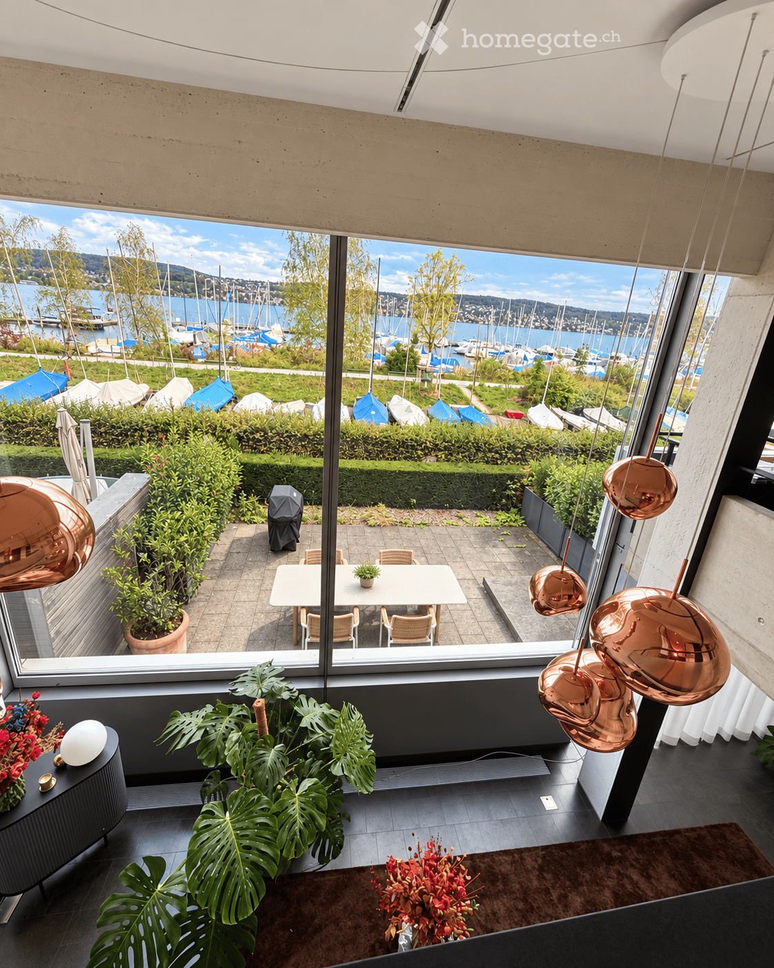 Modern interior with large windows overlooking a waterfront, featuring a dining table, chairs, potted plants, and decorative lighting.