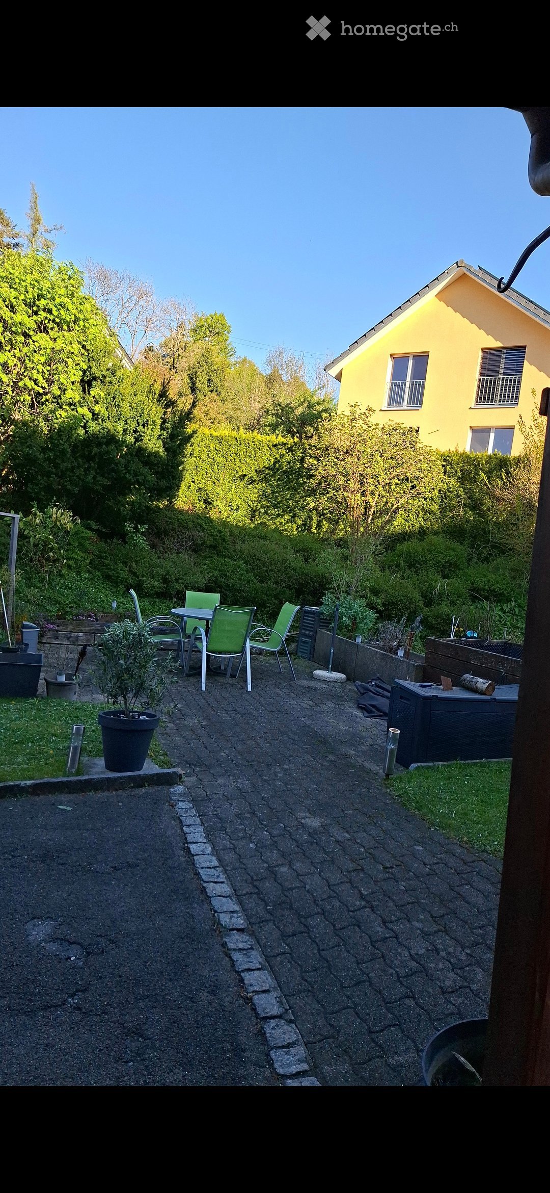 Garden area with a paved pathway, small seating area with a table and chairs, potted plants, and a yellow house in the background.