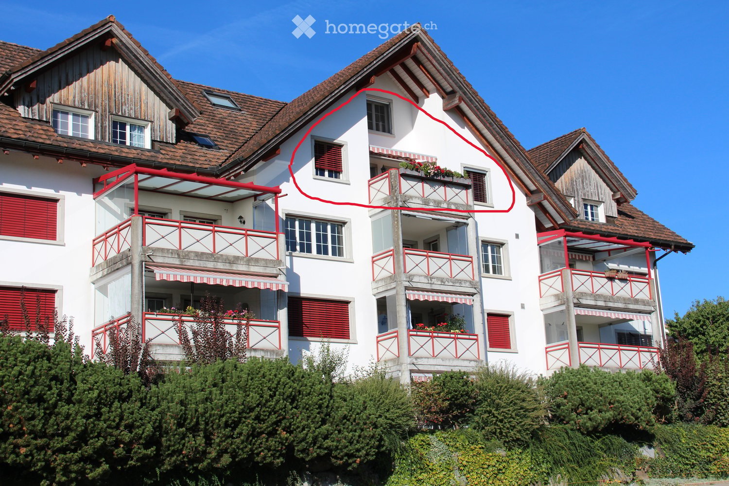 A multi-story residential building with white walls, red shutters, and multiple balconies adorned with flower boxes. The structure features a tiled roof and is surrounded by greenery.
