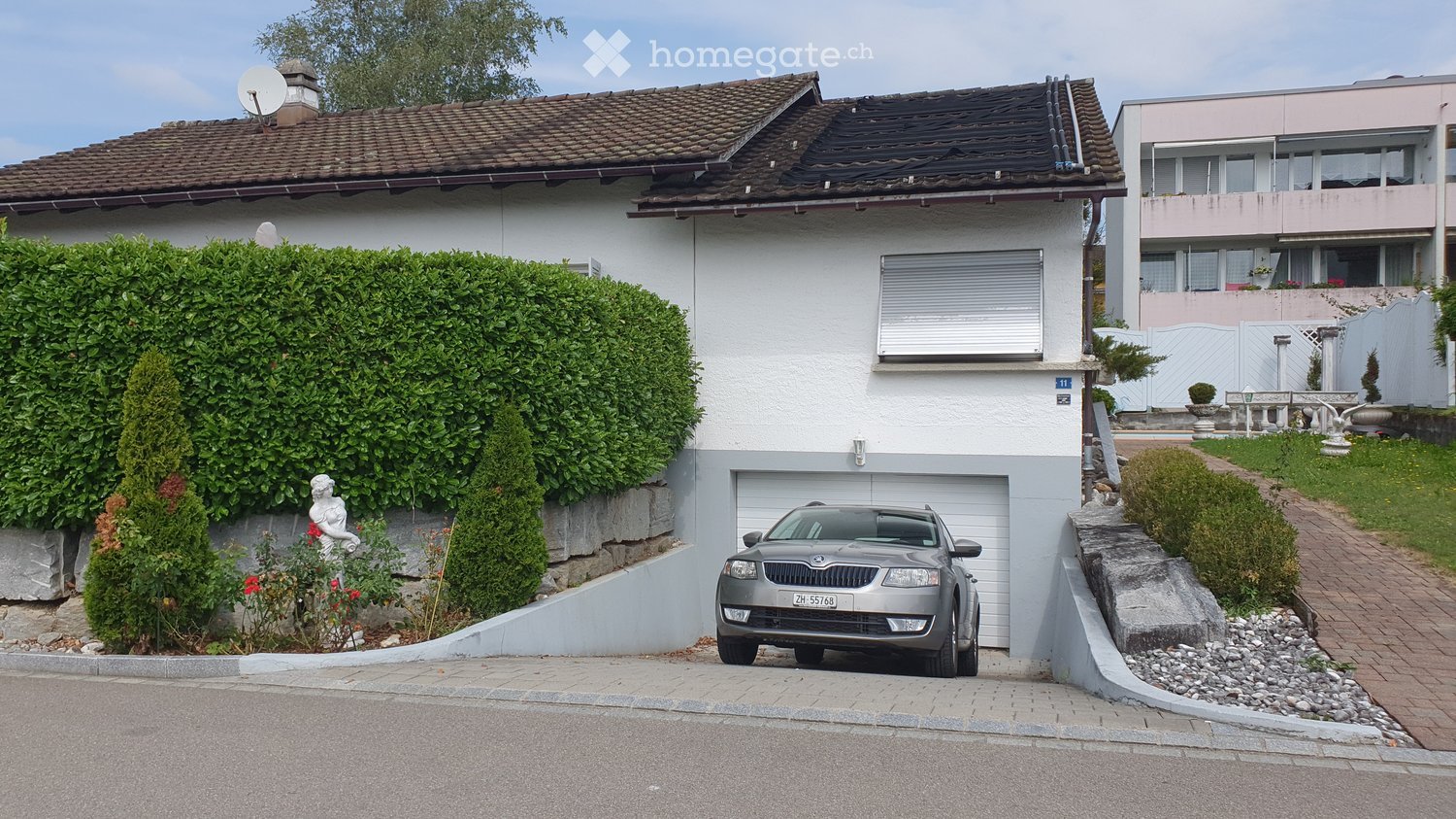 Two-story house with a tiled roof, white exterior, garage, and a parked car in front. The house is surrounded by greenery, including a hedge and small garden area.