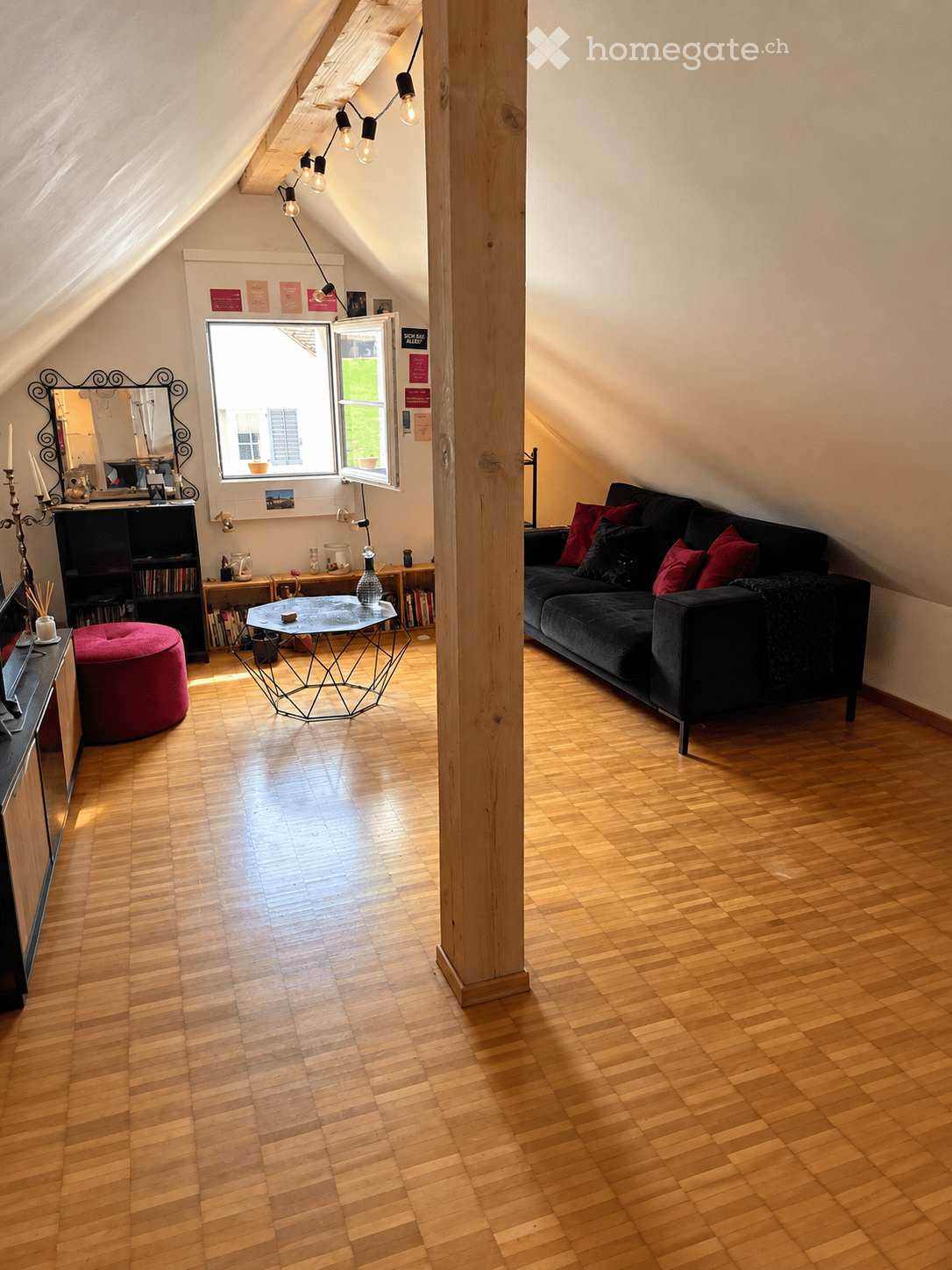 Attic living room with wooden flooring, slanted ceilings, and large windows. Features a dark-colored sofa with red pillows, a geometric coffee table, a red round stool, a bookshelf, and a mirror on the wall.