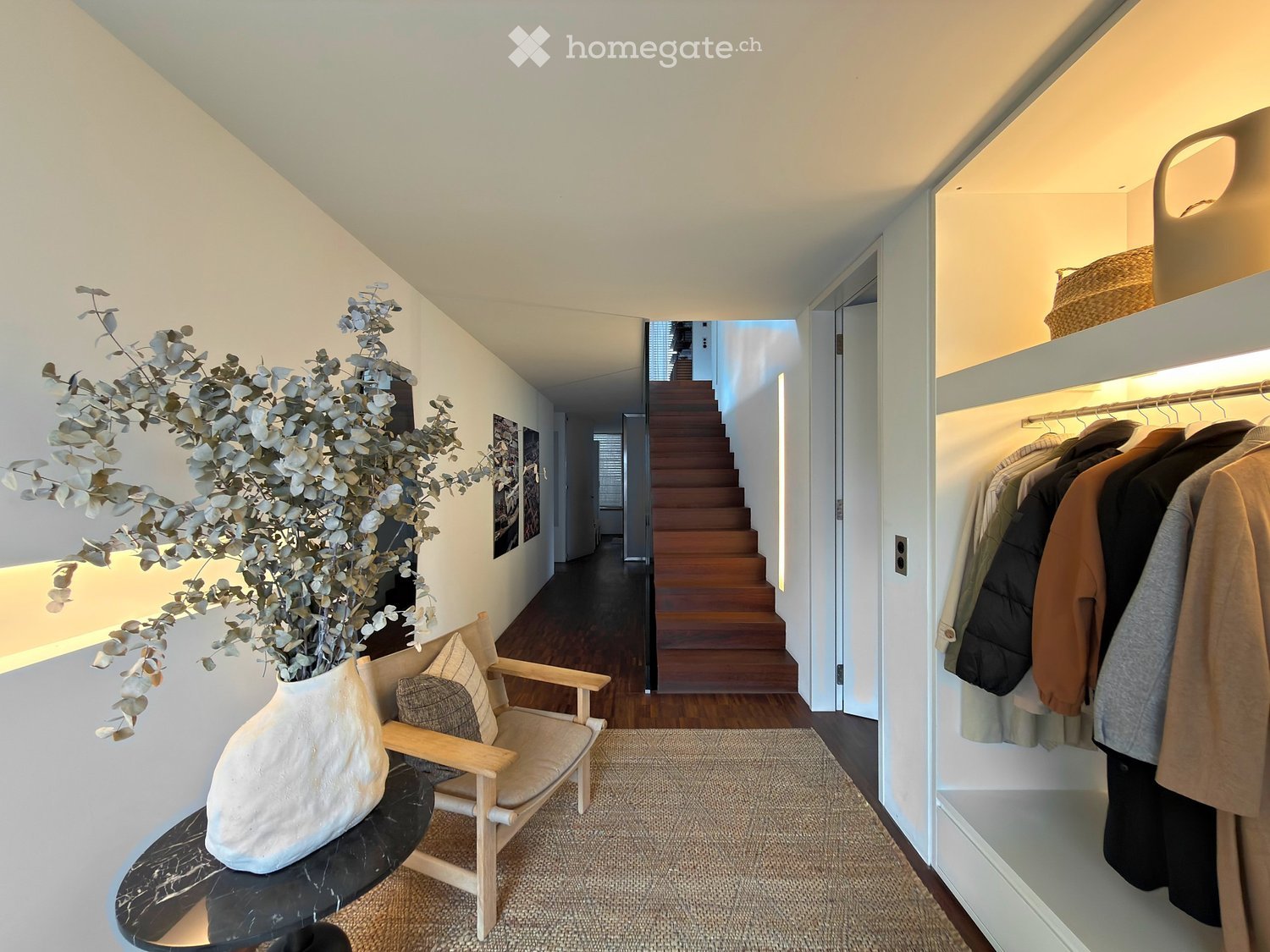 A modern hallway with a wooden staircase, a small seating area with a chair and table, and a coat rack with several jackets. A large vase with dried eucalyptus branches sits on a black marble table.