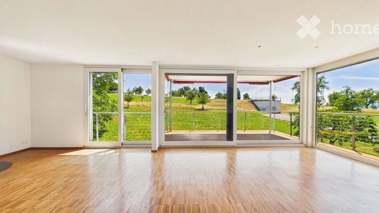 Bright, empty room with large sliding glass doors leading to a patio, wooden flooring, and expansive views of a green landscape.