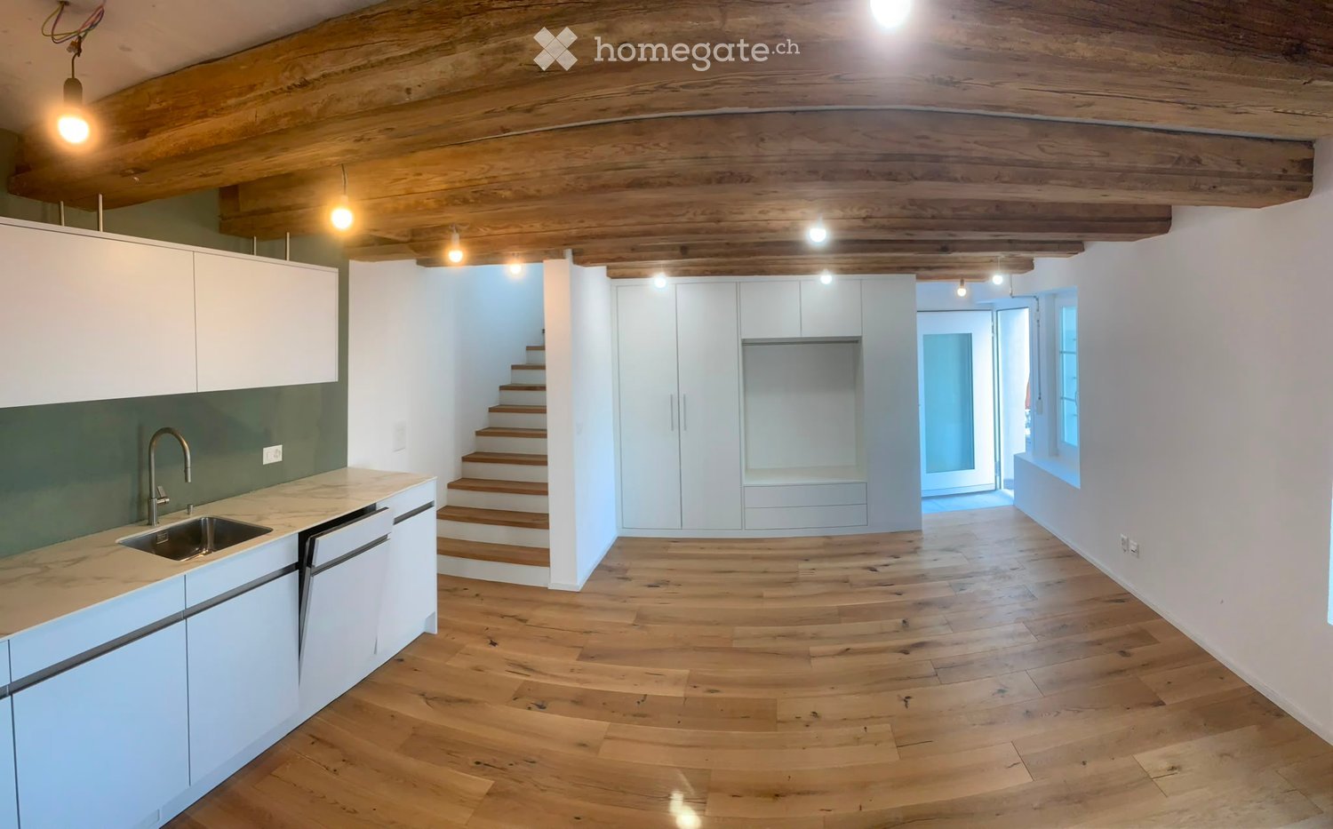 Open-plan space with wooden floors, white cabinetry, a staircase leading to another level, and exposed wooden beams on the ceiling. The area includes a kitchen counter with a sink and a doorway leading to another room.