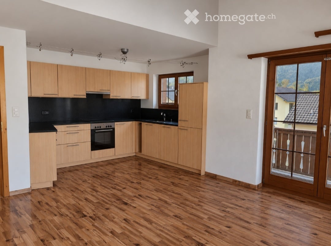 Modern kitchen with light wooden cabinets, black countertops, and a central island. Features include an oven, sink, and ample natural light from large windows and a sliding glass door leading to a balcony.
