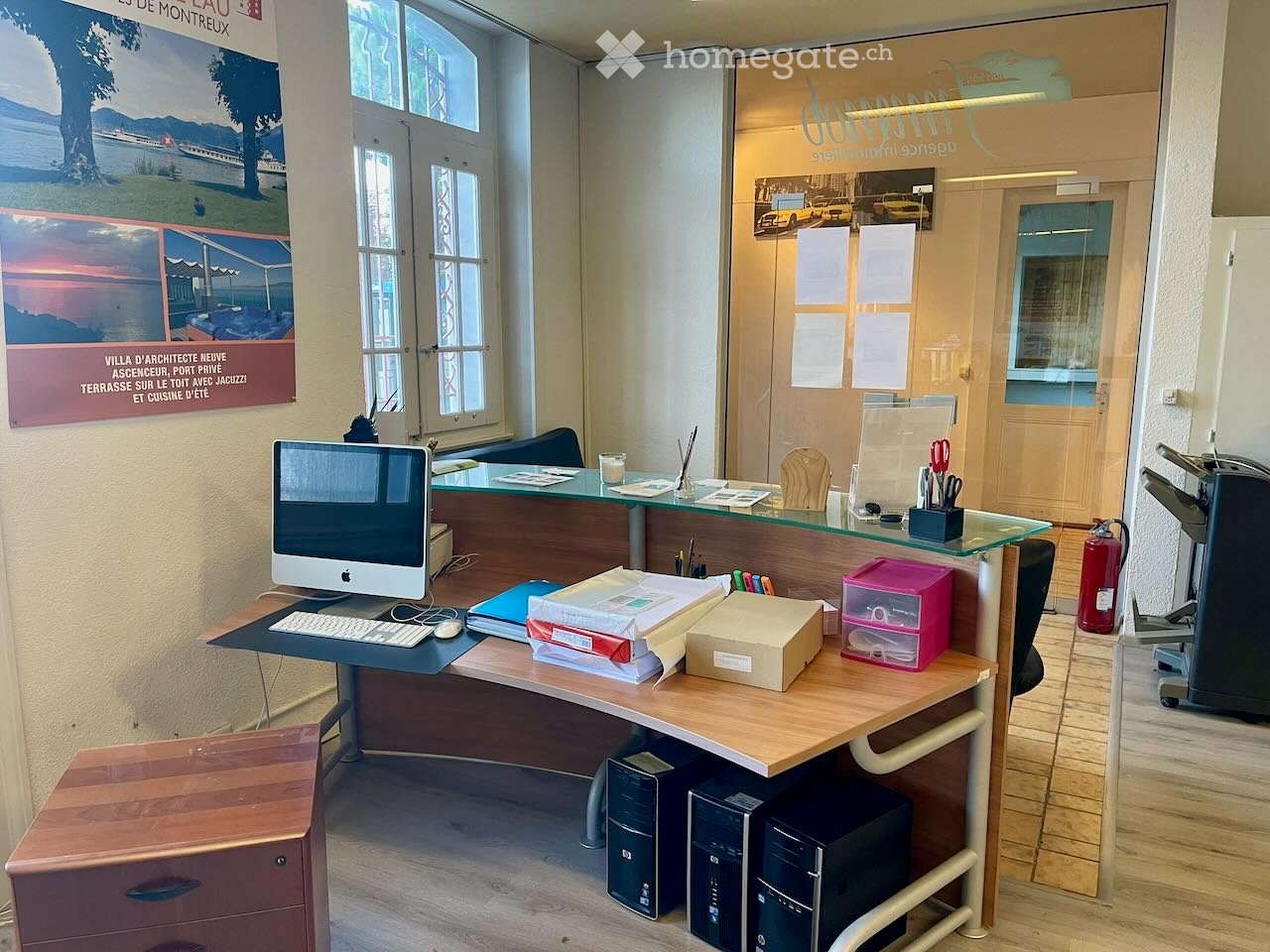 Office space with a wooden desk, computer setup, chairs, filing cabinet, and a glass reception area with office supplies.