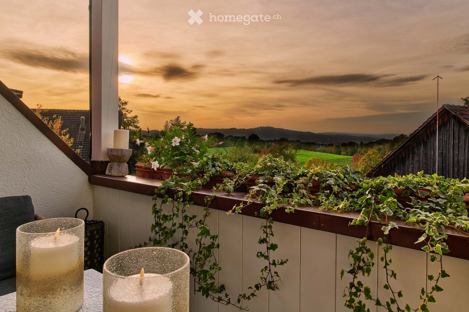 Balcony with potted plants, candles, and a scenic view of a countryside landscape at sunset.