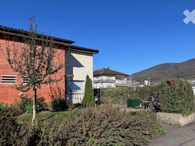 A well-maintained garden area in front of a two-story house with brick and white walls. Features include a small tree, shrubs, a green table, and chairs, with a clear blue sky and distant mountains visible in the background.