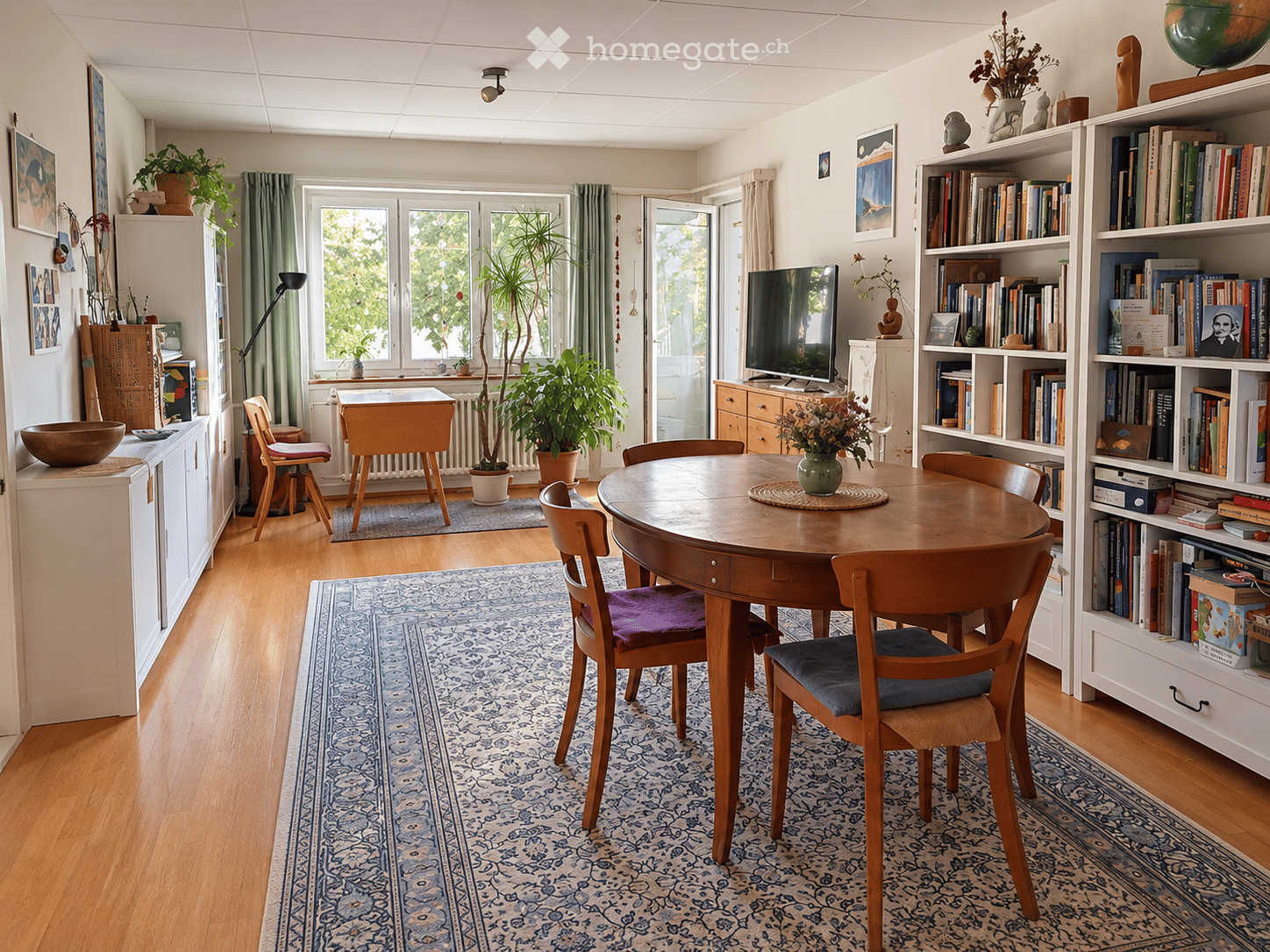 Brightly lit room with wooden flooring, a round wooden dining table surrounded by chairs, a bookshelf filled with books, a television, potted plants, and decorative items.