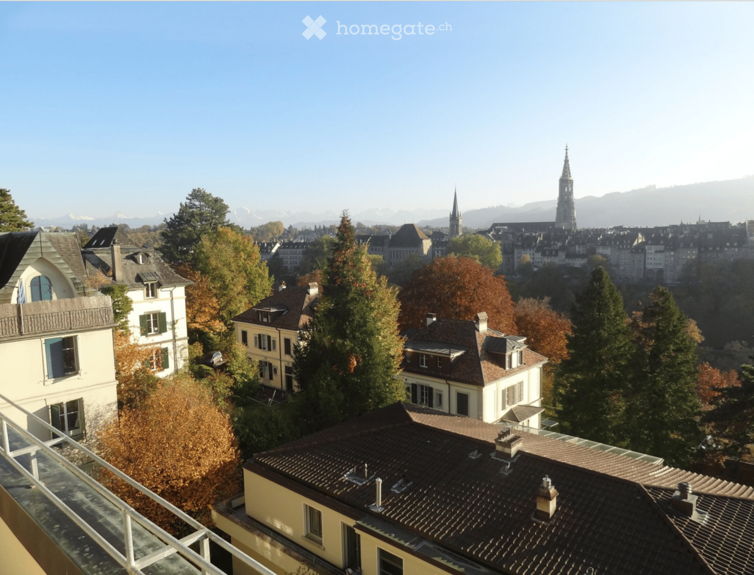 A panoramic view of a cityscape with multiple buildings, trees, and a distant church spire under a clear blue sky.