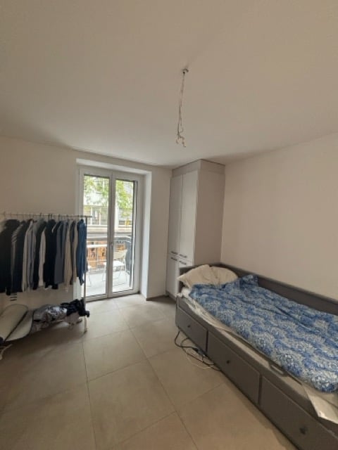 A small bedroom with a single bed, a wardrobe, and a clothing rack. The room has tiled flooring and a large sliding glass door leading to a balcony.