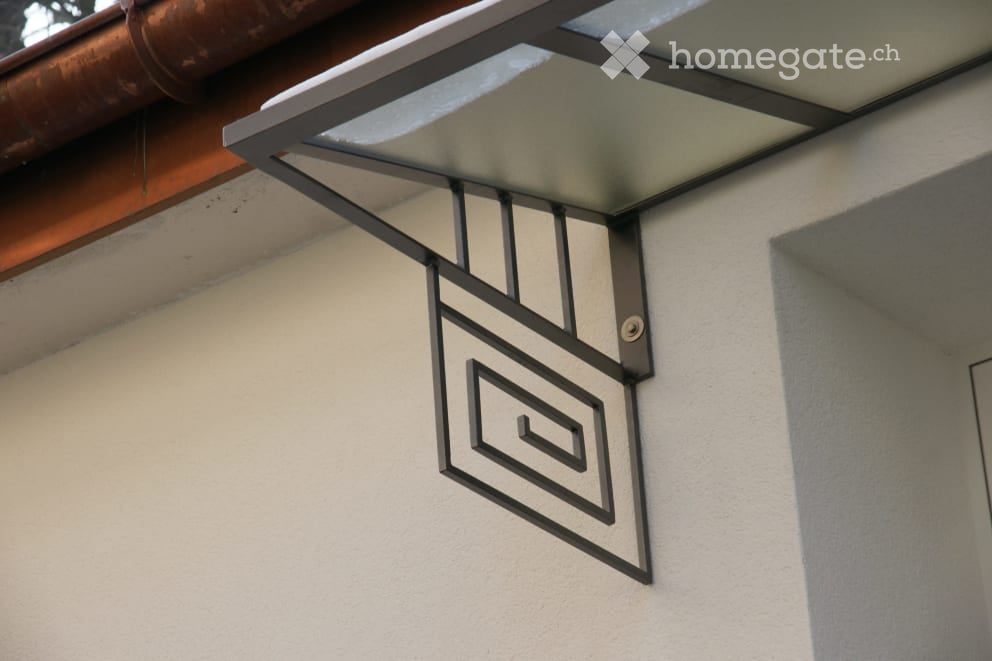 Close-up of a modern building facade featuring a decorative metal sign with a stylized logo and a sleek, glass-covered entrance canopy.