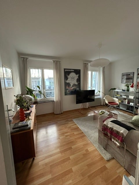 Modern living room with wooden flooring, large windows offering a view, a grey sofa, coffee table, armchair, side table with plants, television, and shelves with books and decor.