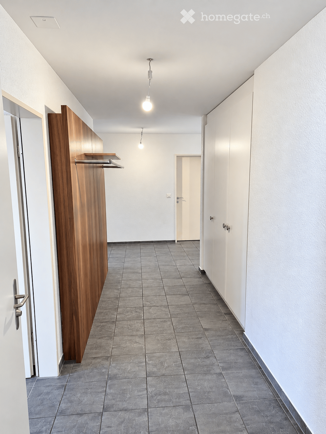 Empty hallway with grey tiled flooring, white walls, and a wooden coat rack on the left side. Two doors are visible, one on each side of the hallway.