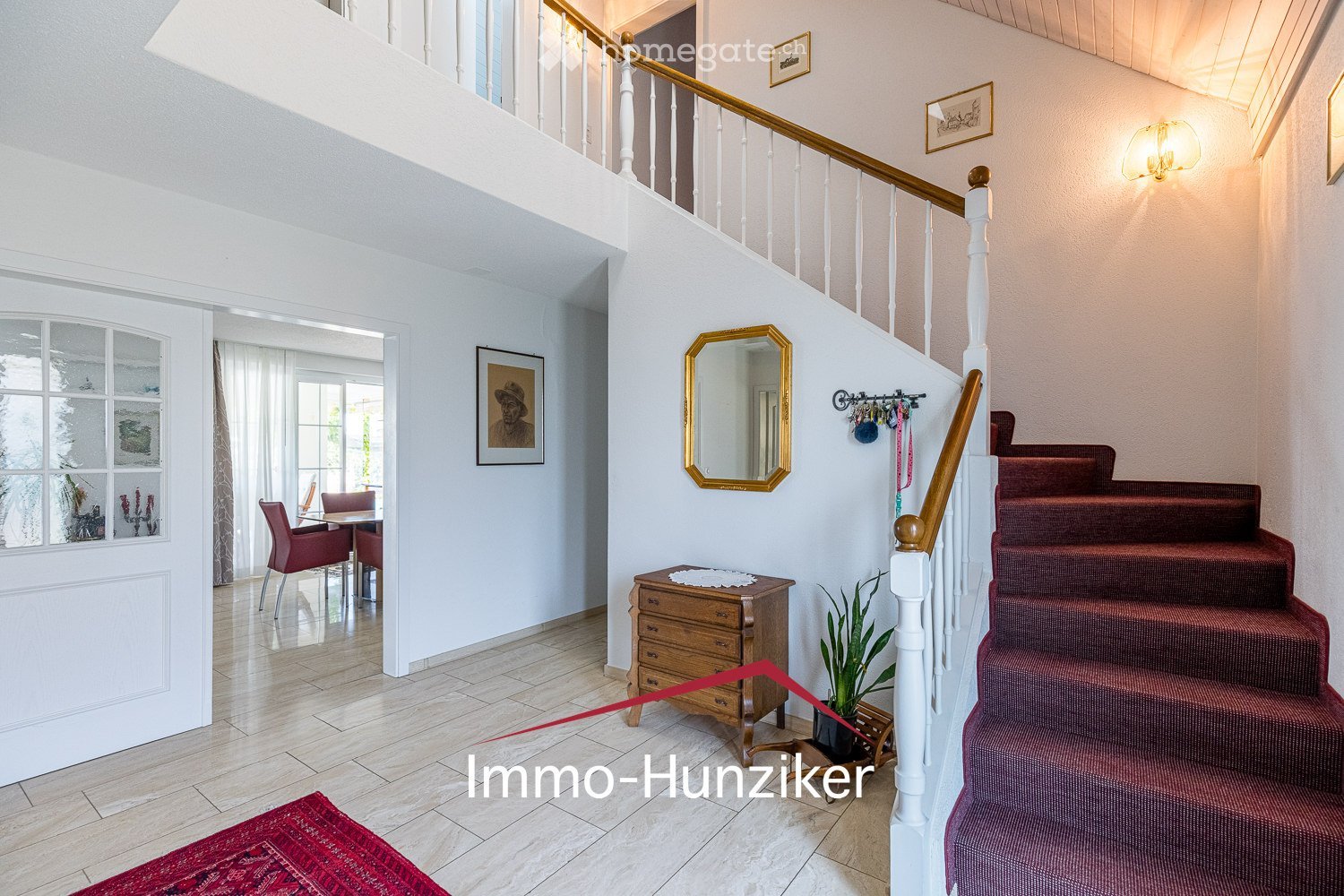 Spacious hallway with a staircase leading to an upper level, wooden railing, red carpeted stairs, a small wooden cabinet, a mirror, and a potted plant. The hallway features white walls, a tiled floor, and several framed pictures.