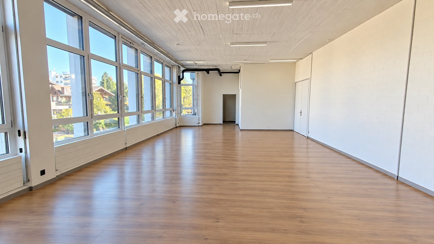 Empty room with wooden flooring, large windows, white walls, and a door leading to another space.