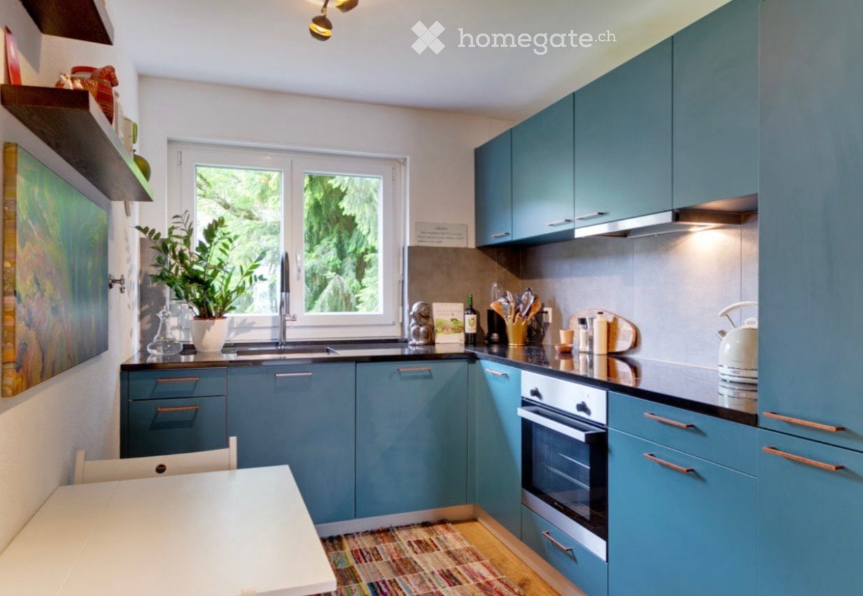 Modern kitchen with blue cabinets, stainless steel appliances, a sink, and a small dining area with a table and chair. A potted plant and a painting add a touch of decor.