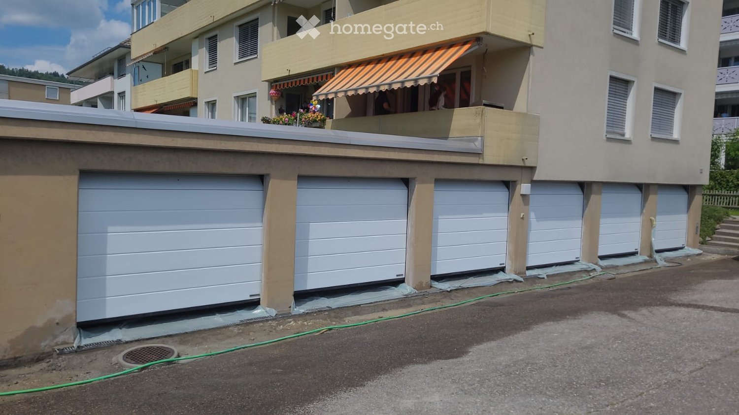 Multi-story residential building with a large garage area featuring multiple garage doors, a small balcony with an awning, and flower boxes.