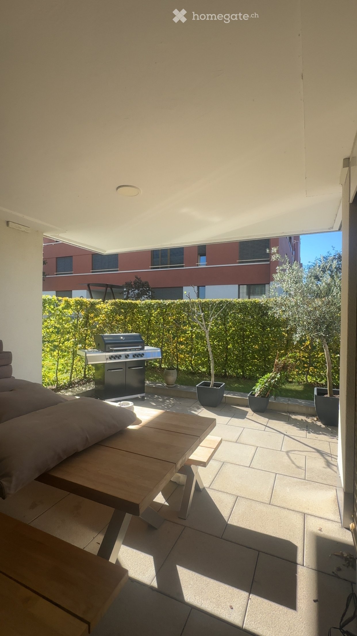 Balcony with a wooden table, sofa, grill, and several potted plants, overlooking a landscaped garden area.
