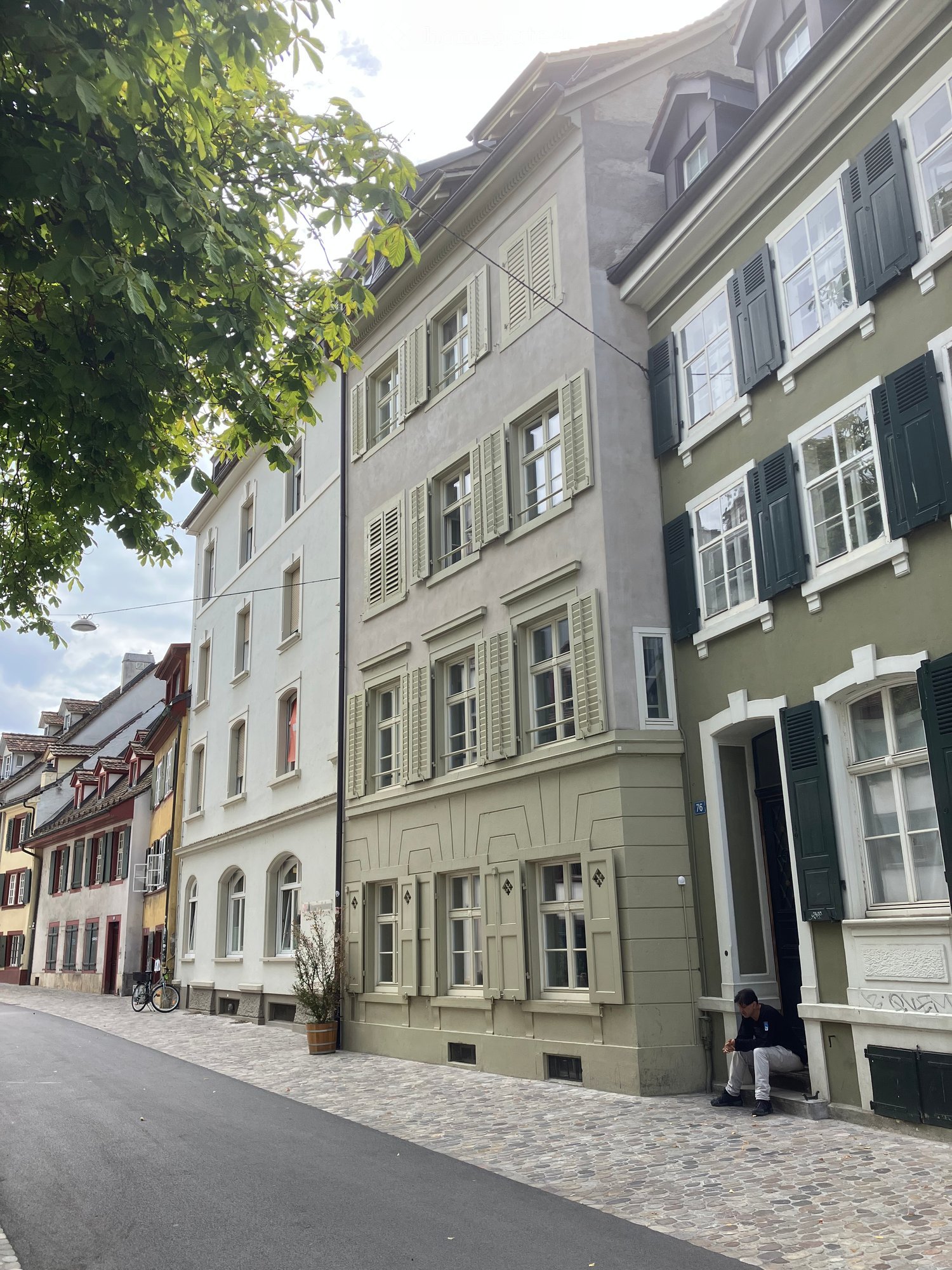 A multi-story residential building with light green and white exterior walls, multiple windows with shutters, and a cobblestone street in front.