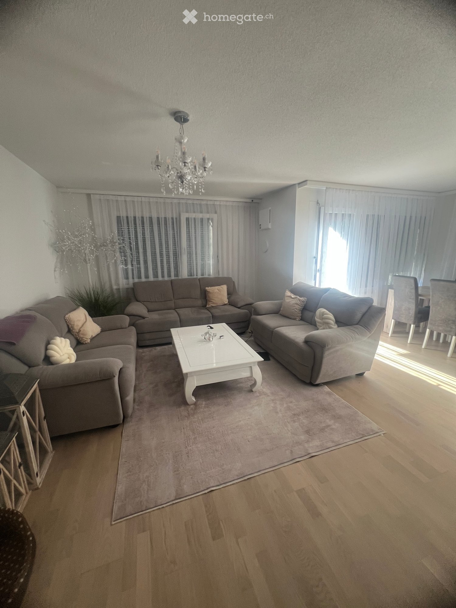 Modern living room with light wooden flooring, a white coffee table, grey sofas, and a chandelier hanging from the ceiling. Large windows with sheer curtains let in natural light.
