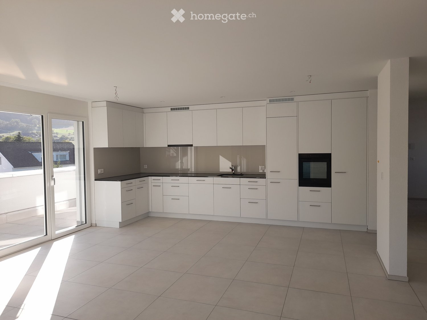 Modern, minimalist kitchen with white cabinets, a central island, and a built-in oven. The space features large windows and a tiled floor.