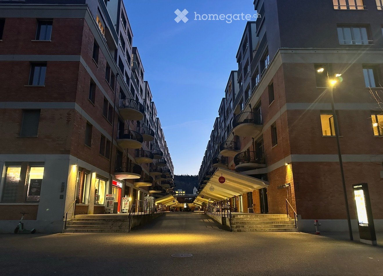 Modern multi-story residential building with balconies and storefronts along a paved walkway.