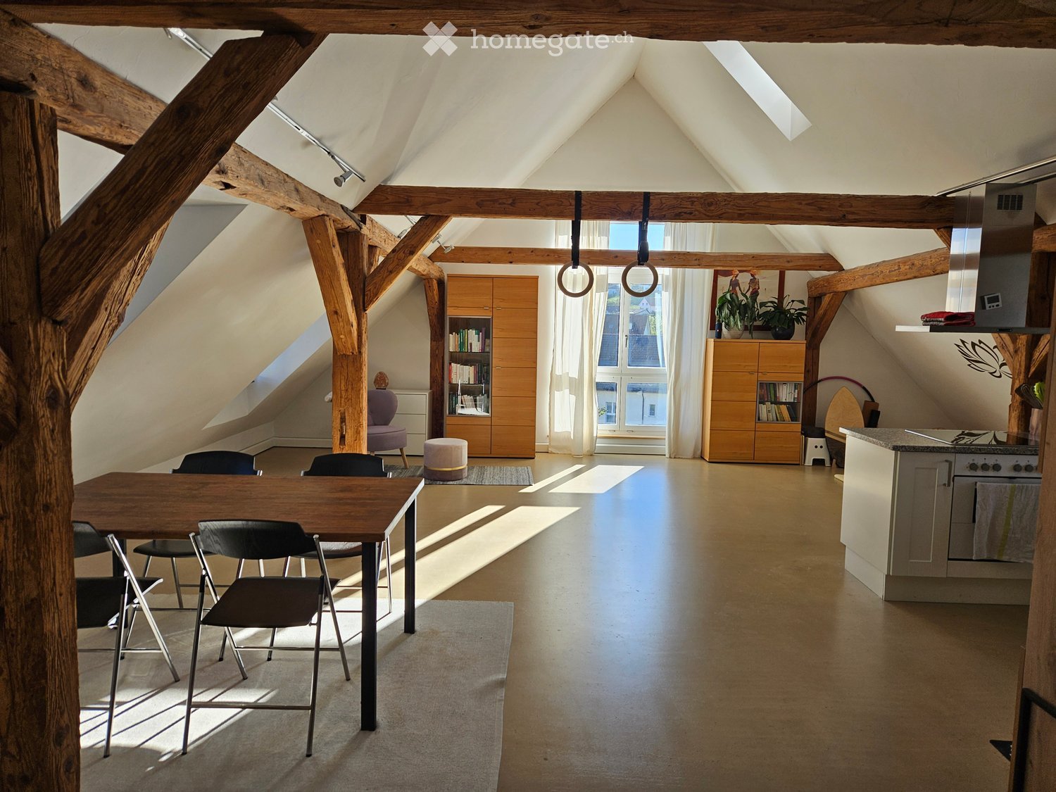 Open-plan attic space with wooden beams, a dining area with a table and chairs, a kitchen area, and built-in wooden cabinets. Natural light streams in from large windows.