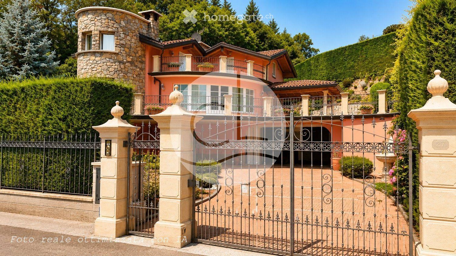 A large, elegant house with a stone tower, pink facade, and multiple balconies surrounded by lush greenery and a decorative iron gate.