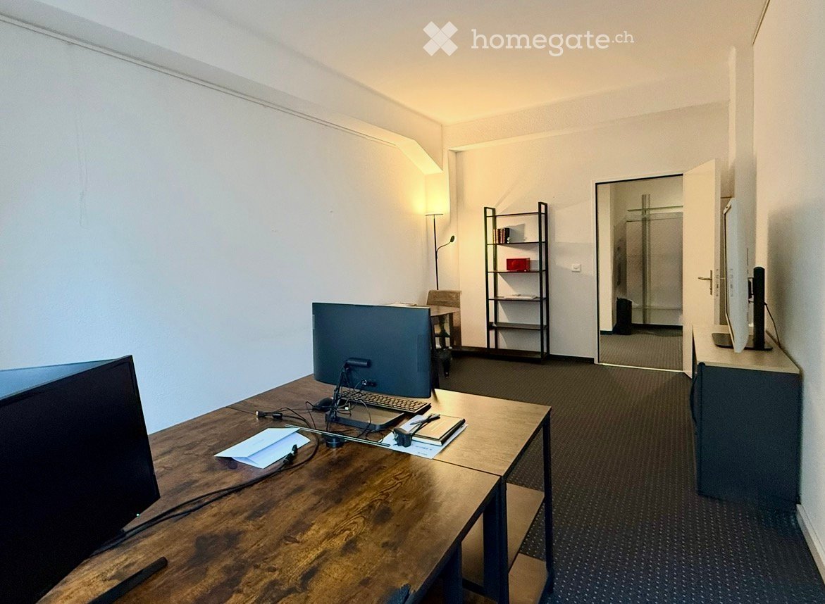 A modern, well-lit office space with a wooden desk, laptop, keyboard, mouse, and a large screen. A small shelf with books and a chair are also visible.