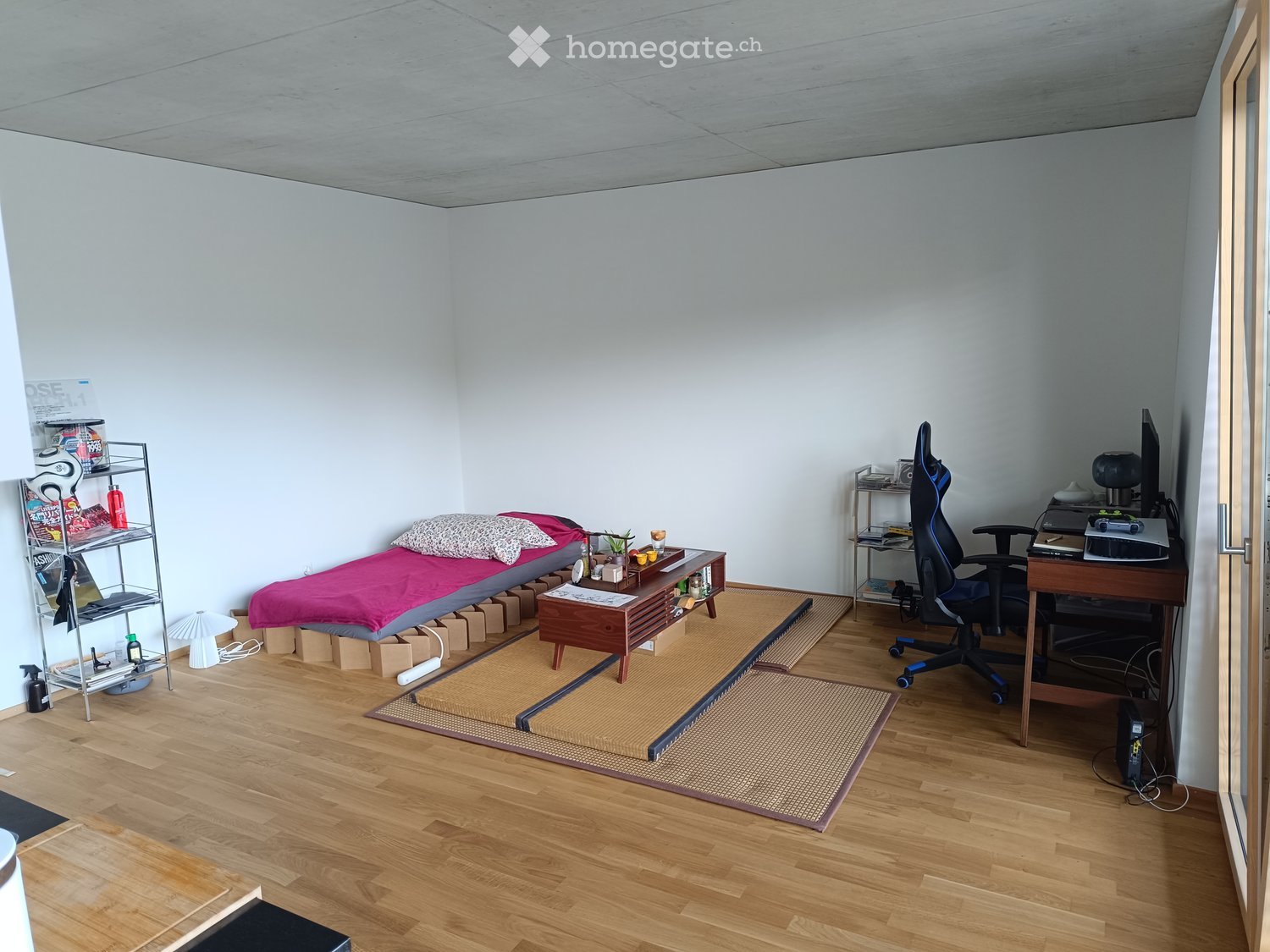 A small, furnished room with a tatami mat area, a bed with pillows, a desk with a chair, and a shelf with various items.