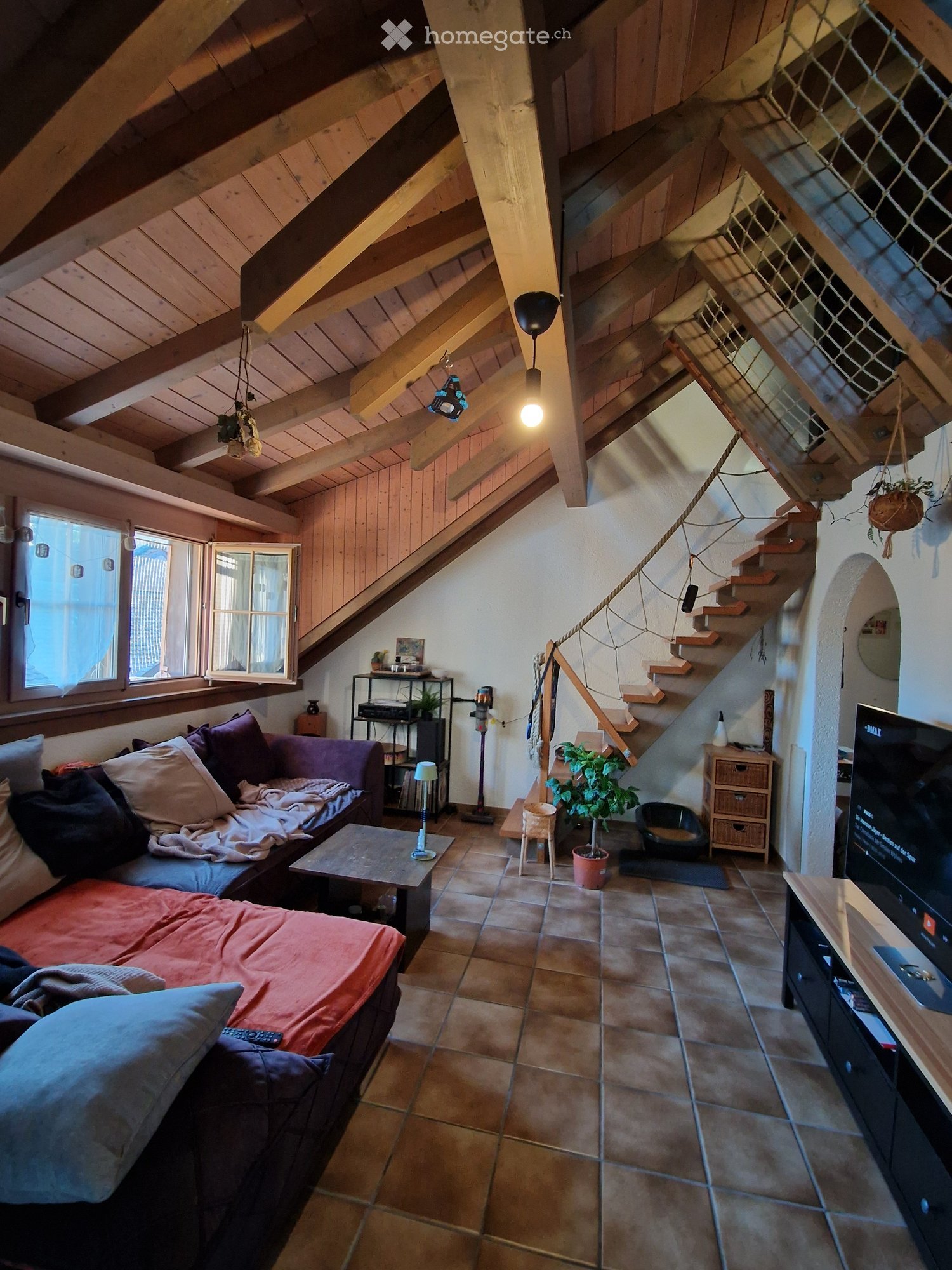 Cozy living room with exposed wooden beams, a large window, a staircase leading upwards, a sofa with pillows, a coffee table, a television, a small table lamp, a potted plant, and a cat bowl.