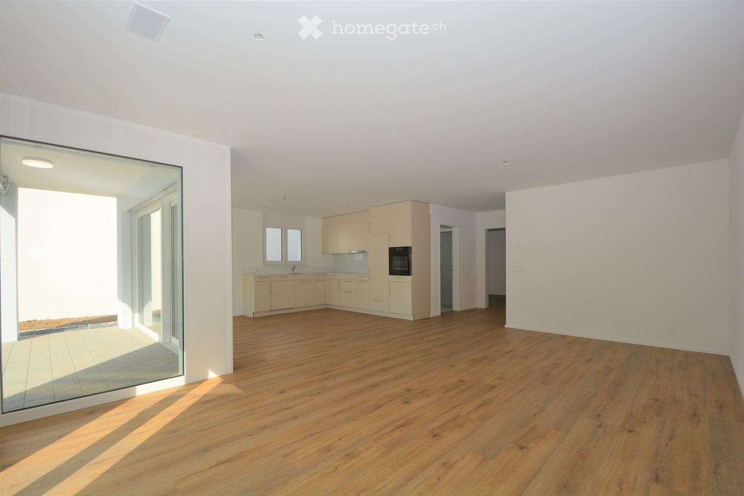 Bright, open-plan living and dining area with wooden flooring, large glass sliding doors leading to a balcony, and a modern kitchen with light-colored cabinets and stainless steel appliances.