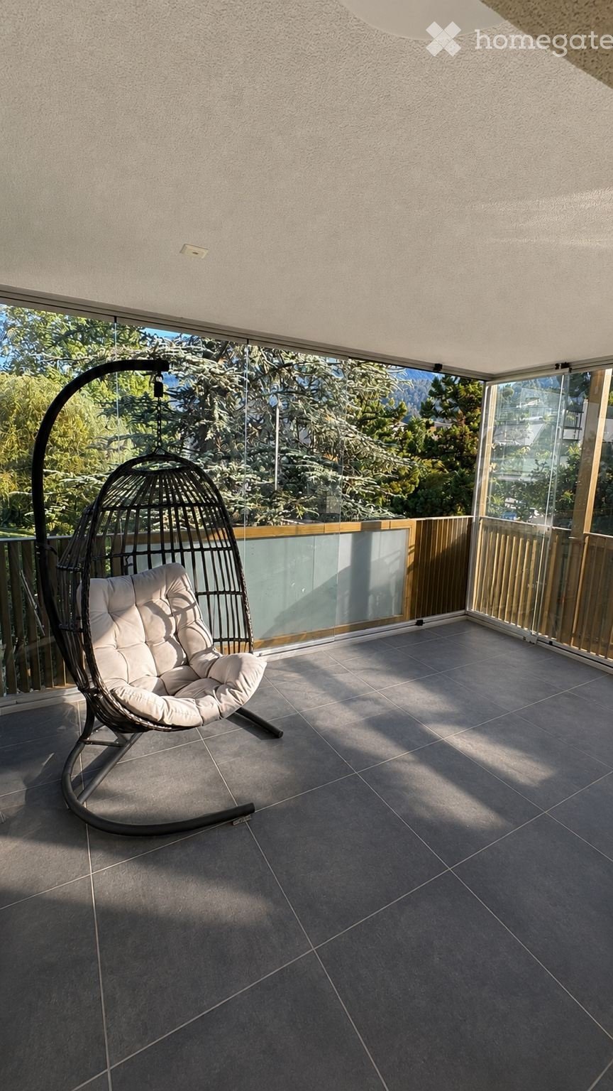 Balcony with tiled floor, glass railing, and a single hanging wicker chair with white cushions.