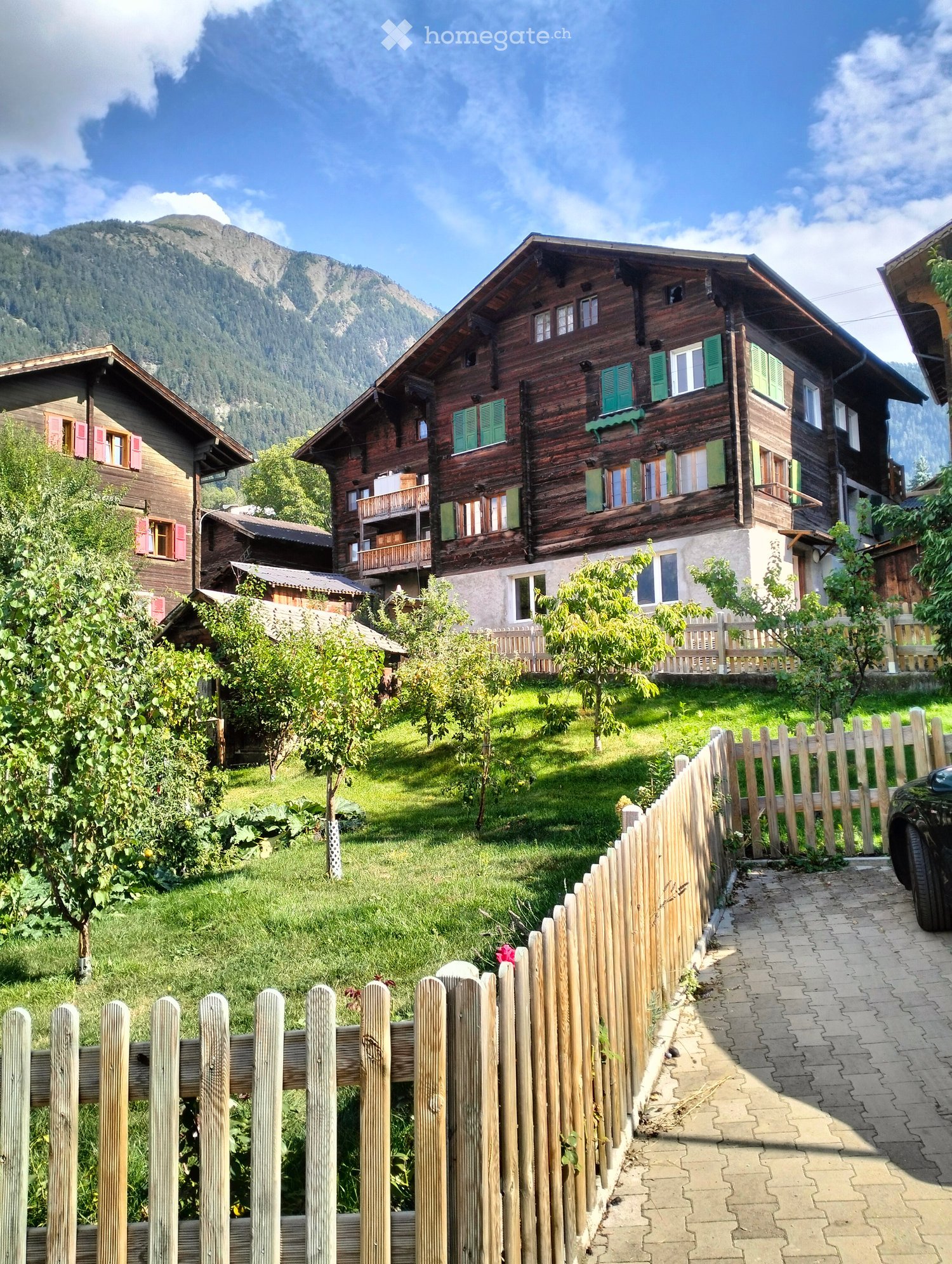 Traditional wooden houses with green shutters and balconies, surrounded by a well-maintained garden and a scenic mountain view.